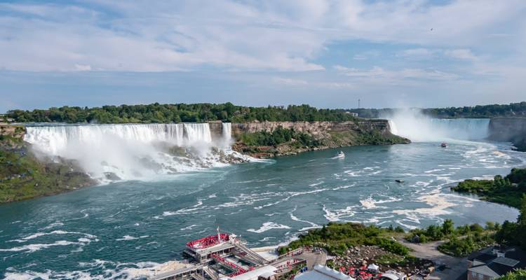 Vue large des chutes du Niagara avec un puissant rideau d'eau blanche et des bateaux d'excursion naviguant sur la rivière tourbillonnante en contrebas.