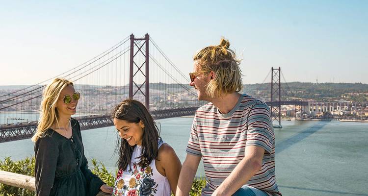 Trois jeunes voyageurs riant à un point de vue panoramique surplombant le pont du 25 Avril qui enjambe le Tage