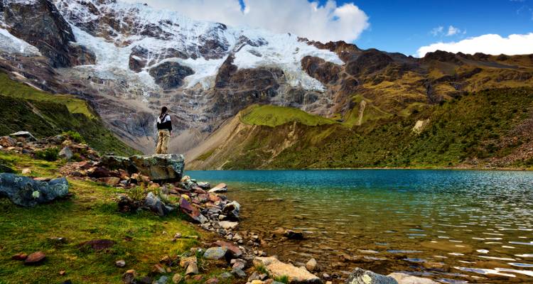 Lone hiker stands on boulder beside turquoise Humantay Lake with snowy Andean peaks towering behind