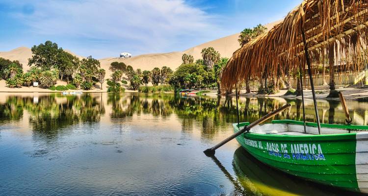 Oasis sereine de Huacachina reflétant les palmiers et les dunes du désert avec une barque verte amarrée sous un auvent de chaume.