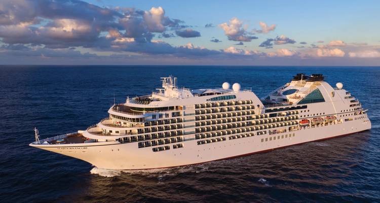 Large white luxury cruise ship sails through calm blue ocean waters at sunset.