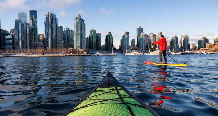 Vue à la première personne en kayak glissant vers un horizon de tours de verre tandis qu'un pratiquant de paddle passe sur une eau calme.