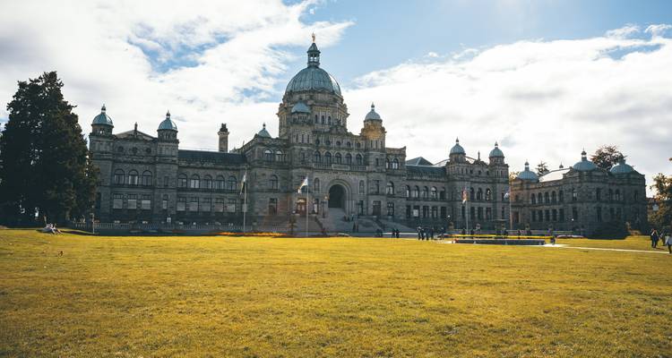 Bâtiment historique du parlement à dôme situé derrière une large pelouse verte par une journée partiellement nuageuse.