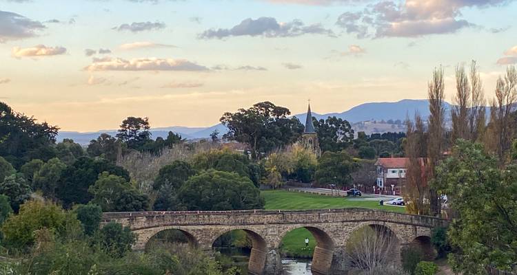 Historic sandstone Richmond Bridge spans a tranquil river with rolling hills and trees at sunset.