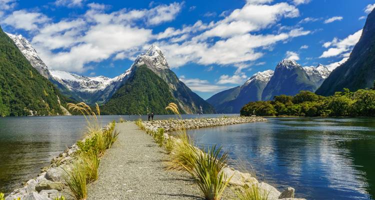 Vue spectaculaire de Mitre Peak dominant Milford Sound avec un sentier dégagé, des pentes luxuriantes et des montagnes enneigées se reflétant dans une eau calme.