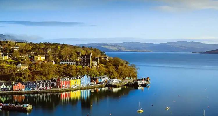 Brightly painted buildings line Tobermory harbour reflecting on calm blue waters with highland hills beyond.