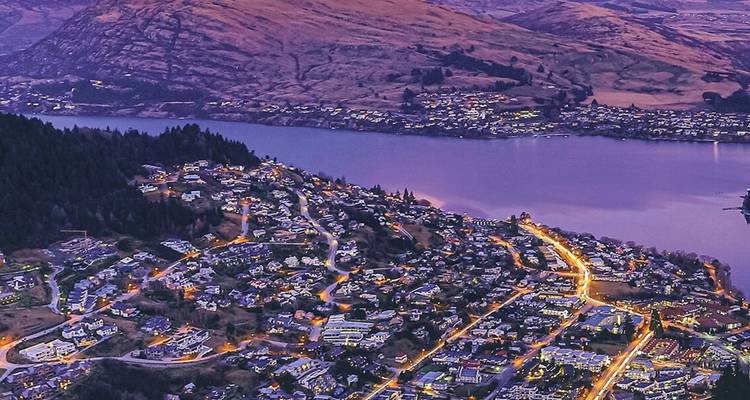 Vue aérienne crépusculaire de Queenstown illuminée au bord du lac Wakatipu et des collines environnantes.