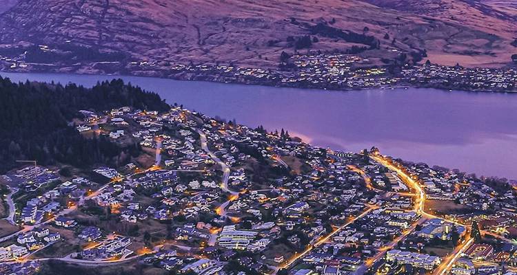 Vue aérienne crépusculaire d'une ville lacustre avec des rues sinueuses illuminées et des montagnes environnantes.