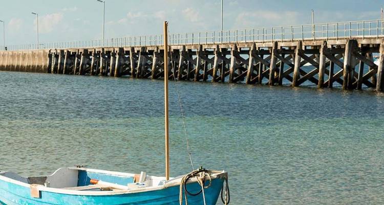 A small blue fishing boat floats beside a long wooden pier stretching over calm coastal waters.