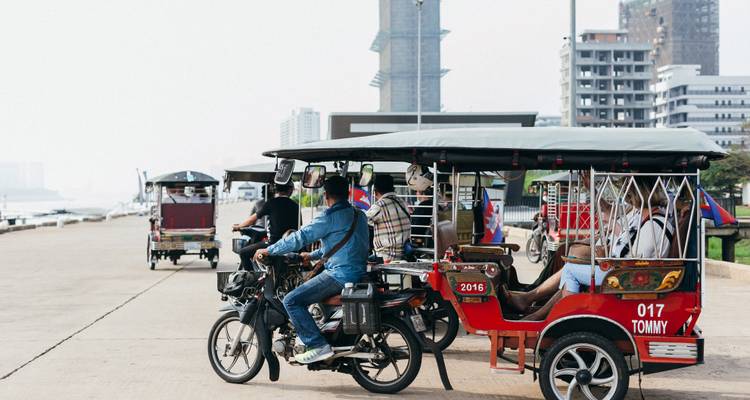 Row of tuk-tuks and motorcycles ready for passengers along a hazy riverside boulevard.