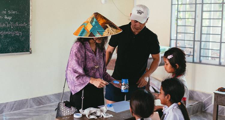 Traveler in conical hat interacts with local children during a classroom visit while another adult observes.