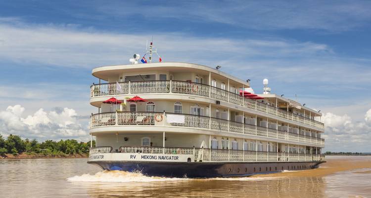 Elegant three-deck river cruiser ‘RV Mekong Navigator’ cutting through brown water under a bright blue sky with scattered clouds.
