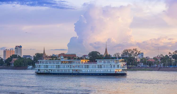 River cruise ship gliding past Phnom Penh skyline at pastel sunset with towering clouds behind the city spires.
