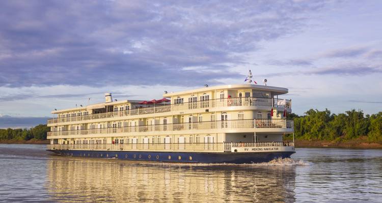 Large white-and-blue river cruise vessel ‘RV Mekong Navigator’ cruising on calm brown water under a cloudy blue sky with green riverbanks.