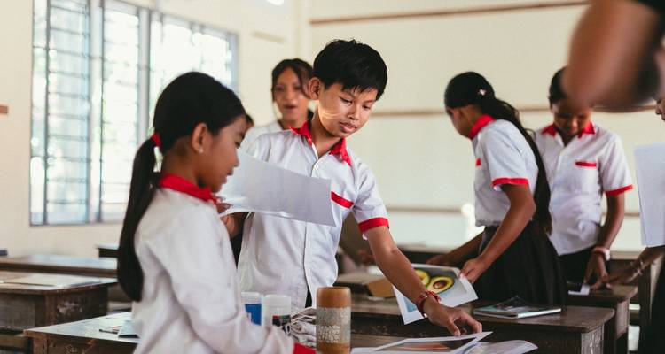 Uniformed schoolchildren work on art projects inside a bright classroom, handling papers and colored materials.