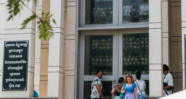 Visitors outside a memorial stupa with glass panels displaying rows of skulls, reading signage and taking photos.