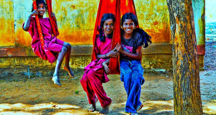Smiling rural Indian children playing on a red cloth swing against a weathered yellow wall.
