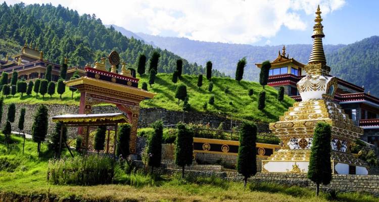 Colorful hillside Buddhist monastery complex with chortens and manicured cypress trees against mountain backdrop.