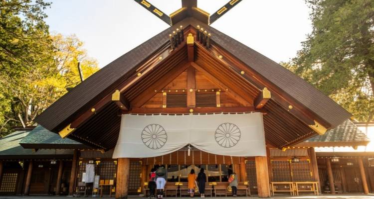 Wooden Shinto shrine with large white curtain and chrysanthemum emblems surrounded by trees in sunlight.