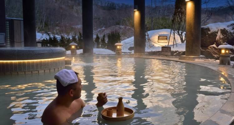 Person relaxes in steaming outdoor onsen pool at dusk with snowy forested hills around.