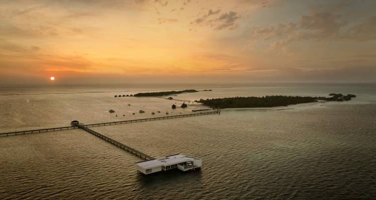 Golden sunset over an over-water villa and long pier stretching to a tiny Maldives island.
