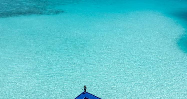 Minimalist aerial shot of a person standing at the end of a blue jetty surrounded by crystal-clear turquoise water.
