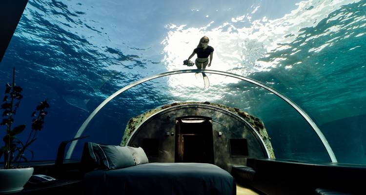 Luxurious underwater bedroom with a glass dome ceiling where a woman swims above, lit by sun rays.