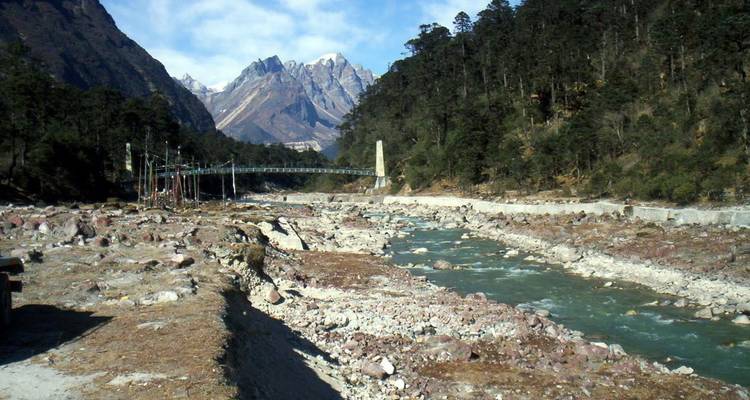 Rocky riverbed with turquoise water flowing through a forested Himalayan valley.