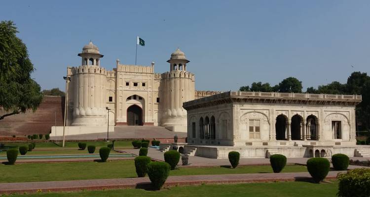 Una puerta histórica de fuerte de piedra arenisca con dos torres y la bandera nacional ondeando arriba, situada contra un cielo azul claro y céspedes cuidados.