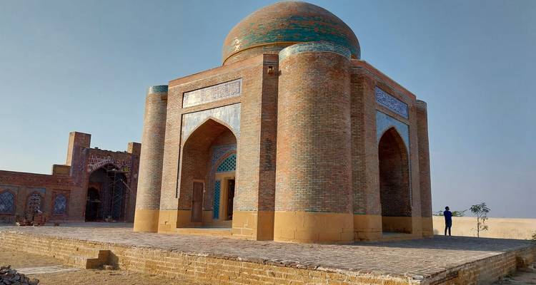 Un mausoleo solitario de ladrillo y teja con una cúpula turquesa se alza en una llanura desértica bajo un cielo despejado.