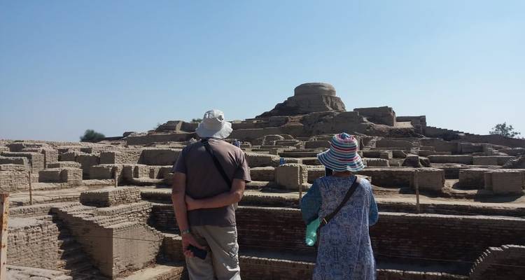 Dos turistas observan extensas ruinas antiguas de ladrillo y una estupa bajo un cielo azul brillante.