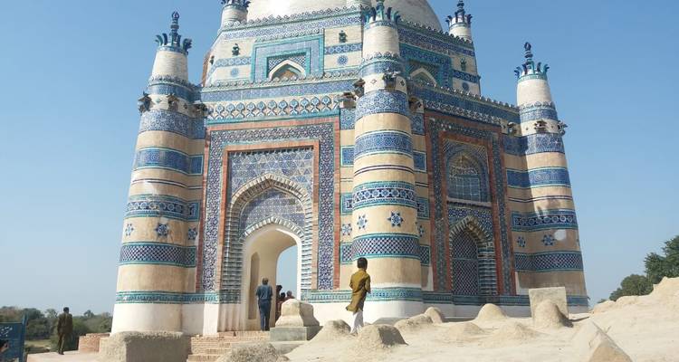 Un mausoleo ricamente decorado con azulejos azules y blancos con patrones intrincados se alza en un paisaje arenoso mientras los visitantes lo contemplan.