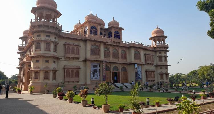Un elegante palacio indo-sarraceno con cúpulas de arenisca rosa y balcones ornamentados se encuentra en un jardín bien cuidado.