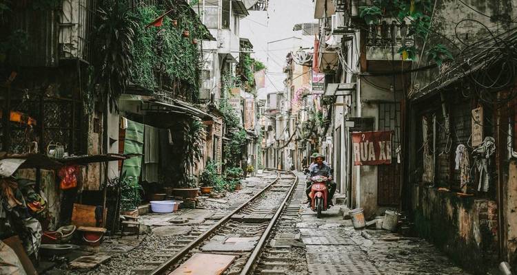 Una motocicleta avanza por las estrechas vías del tren entre casas altas y deterioradas cubiertas de vegetación en la famosa Calle del Tren de Hanói.