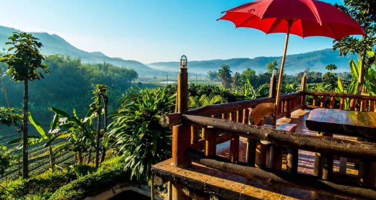 A wooden terrace with a red umbrella overlooks misty green valleys and distant hills under a clear morning sky.