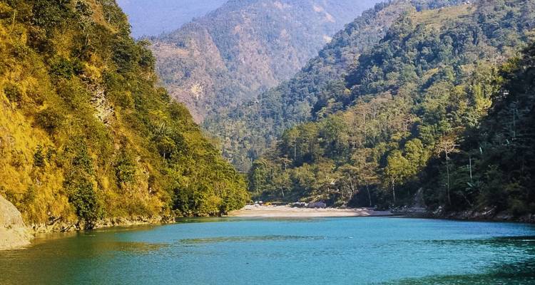 A clear blue river snakes through a steep forested valley framed by lush mountains.