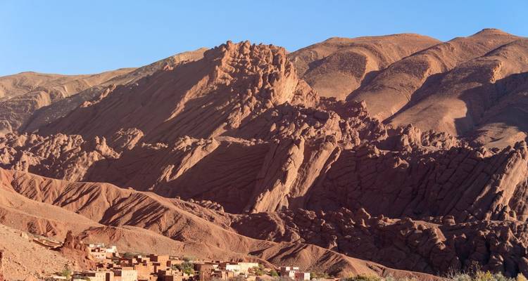 Rugged reddish rock formations tower above a small village at the base of a dramatic valley.