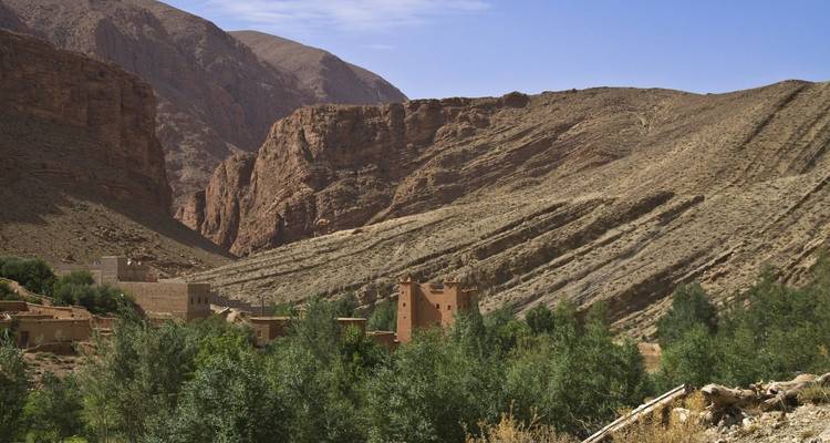 A traditional kasbah sits among green trees with towering rocky cliffs of a gorge behind it.