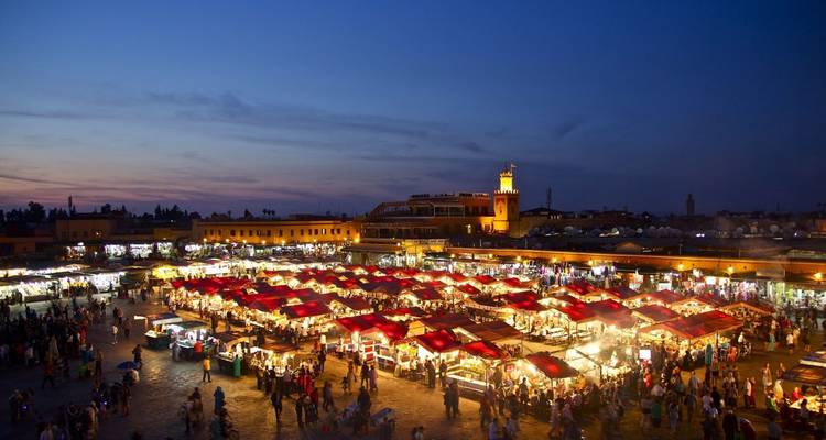 An illuminated night market square with rows of red canopies buzzes with shoppers and food stalls at dusk.