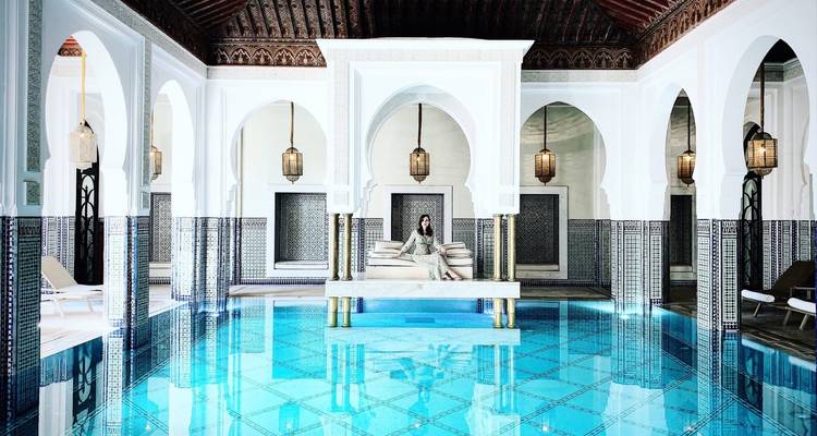 A luxurious riad pool room features turquoise water, white arches and patterned tiles where a woman lounges on cushions.