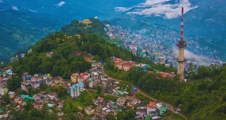 Aerial view of Gangtok town spreading over a forested ridge with a tall communications tower and cloud-shrouded mountains behind.