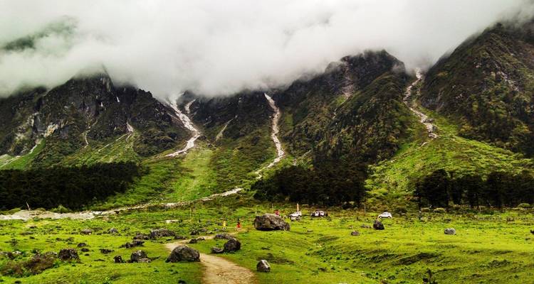 Green alpine valley with scattered boulders, misty clouds clinging to steep slopes and multiple thin waterfalls cascading down.