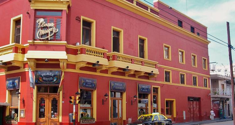 Bâtiment d'angle classique rouge et jaune avec balcons ornés et un taxi qui passe dans une rue ensoleillée de Buenos Aires.