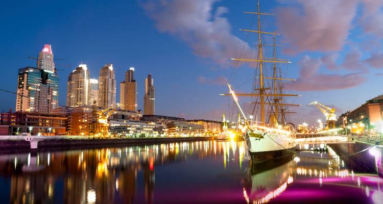 Vue nocturne de l'horizon de Puerto Madero se reflétant sur une eau calme avec un grand voilier historique illuminé au premier plan.