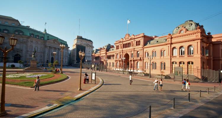 Vue large de la place avec le bâtiment rose de la Casa Rosada sous un ciel bleu dégagé avec des piétons éparpillés.
