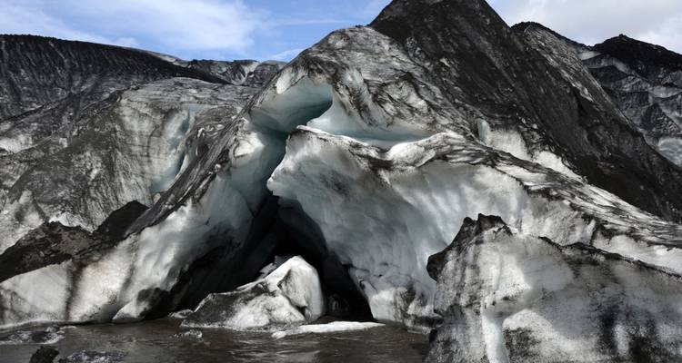 Jagged glacier ice streaked with black ash towers above a meltwater pool, revealing a cavernous opening.