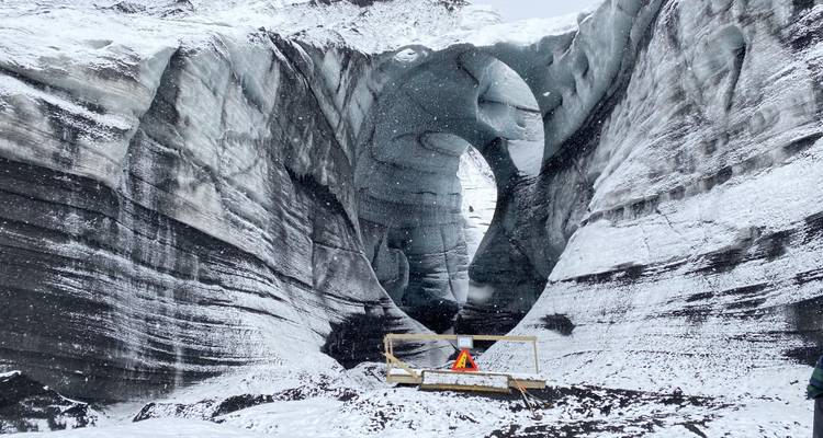 Snow falls around a massive natural ice arch carved into a glacier, surrounded by white, striated walls.