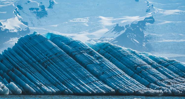 A striking blue iceberg with bold black striations floats against a backdrop of snowy peaks.