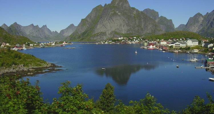 Village pittoresque de fjord avec des montagnes escarpées et déchiquetées se reflétant dans une eau bleue calme sous un ciel dégagé.