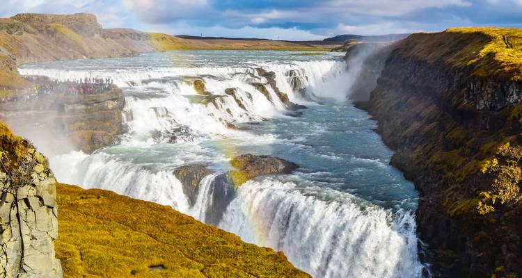 Krachtige cascade van de Gullfoss waterval die in een ruige canyon stort met een zwakke regenboog en kleine toeristen op het uitkijkpunt op de klifrots.